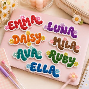 May include: A collection of seven embroidered name patches featuring the names EMMA in red, LUNA in purple, DAISY in orange, MILA in brown, AVA in teal, RUBY in green, and ELLA in blue. Each patch has a wavy font and small flower accents.
