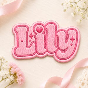 May include: A pink embroidered patch featuring the name Lily in a rounded, bubbly font. The design includes a small heart above the letter i, along with two four-pointed star accents and two small dots, all outlined in a darker shade of pink.