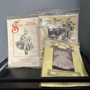 May include: Three sewing patterns for doll clothing in clear plastic sleeves. One pattern is Seeley's Pattern MP40 for Hanna's Dress and Underwear. Another is Ribbons & Roses, and the third is a Doll Club pattern titled White Wedding.