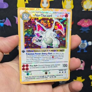 May include: A holographic White Charizard trading card featuring a white dragon illustration, 150 HP, and the Fire Spin attack. The card displays text including Pokémon Power: Energy Burn, weakness, resistance, and retreat cost details.