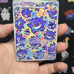 Peut inclure: Une carte à collectionner Gengar avec une finition holographique présentant plusieurs illustrations du personnage violet Gengar sur un fond multicolore vibrant. La carte affiche le texte Gengar, Gnaw-Together et Hollow Dive, ainsi que des statistiques.