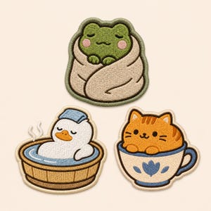 May include: Three embroidered patches featuring a green frog wrapped in a beige blanket, a white duck with a blue towel in a wooden tub, and an orange tabby cat sitting inside a white teacup with blue floral patterns and a blue handle.