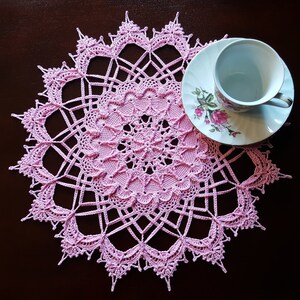 May include: A delicate pink crocheted doily with a floral design. The doily is laid flat on a dark brown surface.