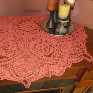 Puede incluir: Un camino de mesa de ganchillo de color coral con un delicado patrón de encaje. El camino de mesa está drapeado sobre una superficie de madera con tres candelabros y velas.