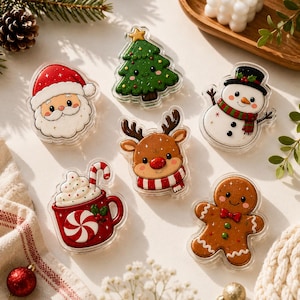 May include: A collection of six acrylic Christmas-themed pins featuring a smiling Santa Claus, a green pine tree with a star, a snowman with a black hat, a reindeer with a red nose, a red mug of hot cocoa with a candy cane, and a gingerbread person.