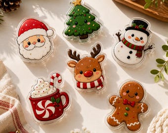 May include: A collection of six acrylic Christmas-themed pins featuring a smiling Santa Claus, a green pine tree with a star, a snowman with a black hat, a reindeer with a red nose, a red mug of hot cocoa with a candy cane, and a gingerbread person.