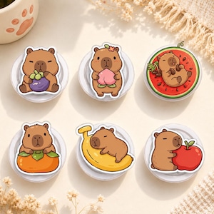 Könnte beinhalten: Ein Set aus sechs runden Handyhaltern mit Cartoon-Wasserschweinen. Die Motive zeigen ein Wasserschwein mit einer Mangostane, einem Pfirsich, in einer Wassermelone, mit einer Kaki, auf einer Banane und einen roten Apfel umarmend. Jeder Halter ist weiß.