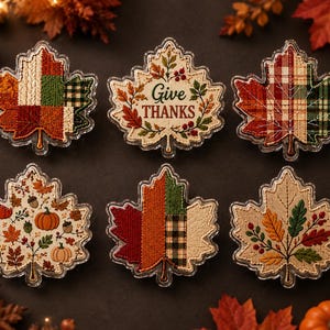 May include: Six leaf-shaped ornaments featuring embroidered autumn designs. One displays the text Give THANKS surrounded by foliage. Others feature patchwork patterns, pumpkins, acorns, and colorful fall leaves in shades of orange, red, and green.