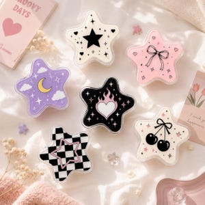 May include: Six star-shaped containers with various designs: a black star, a pink bow, a yellow crescent moon with clouds, a flaming heart, a black and white checkerboard pattern with a pink star, and two black cherries with pink stars.