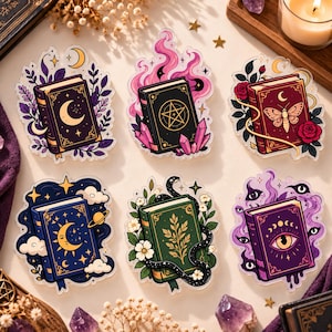 Peut inclure: Un ensemble de six autocollants décoratifs en acrylique représentant des livres illustrés. Les motifs incluent un livre lune violet, un livre pentagramme noir avec cristaux roses, un livre papillon et rose rouge, un livre céleste bleu, un livre serpent vert et un livre œil violet.