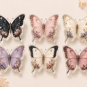 以下が含まれることがあります： 2列に並べられた6つの蝶の装飾品。各蝶は、花柄のアクセント、星や月などの天体のモチーフ、黒、ピンク、クリーム、紫などの多様なカラーパレットを備えた複雑な羽の模様が特徴です。