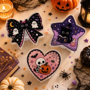 Op de afbeelding: Drie decoratieve telefoonhouders met Halloween-illustraties: een zwarte strik met spoken en sterren, een paarse stervorm met een zwarte kat die een boek leest, en een roze hartvorm met een skelet dat een pompoen vasthoudt.