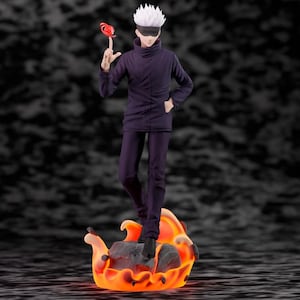 May include: A standing anime character figurine with spiky white hair and a black blindfold, wearing a dark purple high-collar jacket and matching trousers. The figure stands on a base shaped like a rock surrounded by orange and yellow flames.