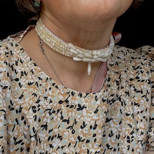 Puede incluir: Un collar tipo gargantilla de color crema con hileras de pequeñas cuentas y perlas alargadas en forma de arroz con una perla central colgante, usado con una blusa de manga corta con un patrón floral abstracto en tonos beige, negro y blanco.