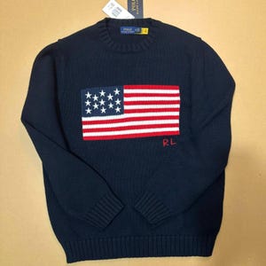 Könnte beinhalten: Ein dunkelblauer Langarmpullover mit Rundhalsausschnitt und einem gestrickten amerikanischen Flaggendesign auf der Vorderseite. Die Flagge zeigt weiße Sterne auf blauem Grund sowie rote und weiße Streifen. Die Buchstaben RL sind rot eingestickt.