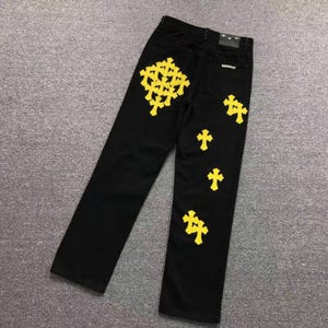 May include: Black denim jeans featuring a series of bright yellow cross-shaped patches. The design includes a large cluster of crosses on the upper left leg and several individual crosses scattered down the right leg, with a black leather patch.