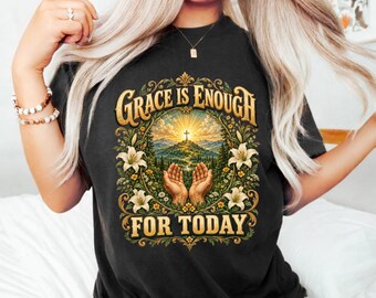May include: A black t-shirt featuring a graphic print with the text "GRACE IS ENOUGH FOR TODAY". The design includes a central illustration of two hands held in prayer, a mountain landscape with a cross, and a border of white lilies.