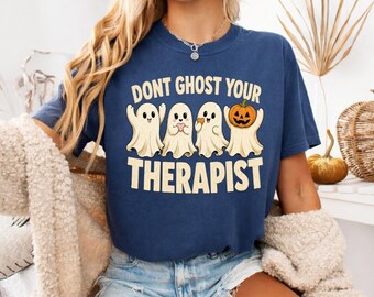 May include: A navy blue short-sleeve t-shirt featuring a graphic of four white ghosts and the text DONT GHOST YOUR THERAPIST in cream-coloured letters. One ghost holds a small orange pumpkin. The shirt has a crew neckline and a relaxed fit.