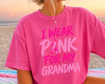 May include: A bright pink short-sleeved t-shirt featuring the text I WEAR PINK FOR MY GRANDMA in a bold, distressed font. A pink awareness ribbon graphic is positioned in the centre of the word PINK, replacing the letter I.
