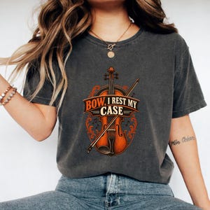 Könnte beinhalten: Ein anthrazitfarbenes Kurzarm-T-Shirt mit einer Grafik einer braunen Geige und eines Bogens. In der Mitte befindet sich ein orangefarbenes Banner mit dem Text "BOW, I REST MY CASE" in fetten schwarzen Buchstaben, umgeben von orangefarbenen Verzierungen.