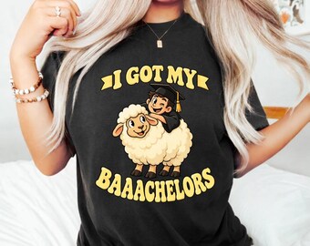 May include: A black t-shirt featuring a graphic of a cartoon person wearing a graduation cap and riding a fluffy white sheep. The text above and below the illustration reads I GOT MY BAAACHELORS in bold yellow capital letters.