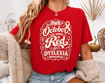 May include: A red short-sleeved t-shirt featuring a white graphic design with the text In October we wear Red for DYSLEXIA AWARENESS, accompanied by illustrations of an open book, a ribbon, a lightbulb, a heart, and an apple.