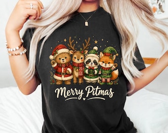 May include: A black t-shirt featuring a graphic print of a bear, a deer, a raccoon, and a fox dressed in Christmas attire. The bear wears a Santa suit, the deer a scarf, the raccoon a green hat, and the fox an elf outfit. The text Merry Pitmas is below.