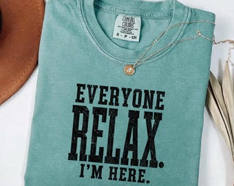 May include: A seafoam green short-sleeve t-shirt with a crew neckline. The shirt features black block lettering that reads "EVERYONE RELAX. I'M HERE." A silver-toned chain necklace with a small circular pendant rests on the collar of the shirt.