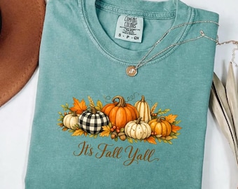 May include: A teal crew neck t-shirt with a Comfort Colors label featuring a graphic of various pumpkins, including one with a black and white buffalo check pattern, surrounded by autumn leaves and the text It's Fall Yall.