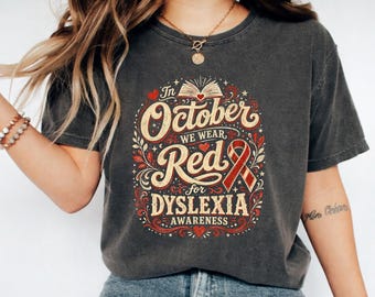 May include: A charcoal grey short-sleeve t-shirt featuring a graphic design with the text In October We Wear Red for Dyslexia Awareness. The design includes an open book, red hearts, decorative leaves, and a red awareness ribbon in a vintage style.