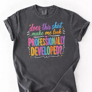 May include: A charcoal grey short-sleeve t-shirt featuring the colourful text "does this shirt make me look PROFESSIONALLY DEVELOPED?" in a mix of pink, blue, yellow, and green letters, accented with small star and line graphics.
