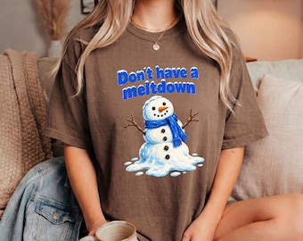 May include: A brown short-sleeve t-shirt featuring a graphic of a melting snowman wearing a blue scarf and the text "Don't have a meltdown" in blue letters. The snowman has stick arms, a carrot nose, and black coal buttons on its body.