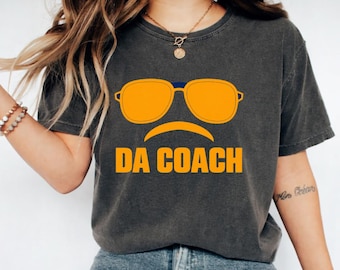 May include: A charcoal grey short-sleeve t-shirt featuring a graphic of orange aviator sunglasses above a curved orange line and the text DA COACH in bold orange capital letters centred on the chest.