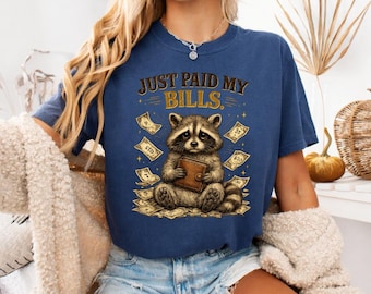 May include: A navy blue short-sleeve t-shirt featuring a graphic of a raccoon holding a brown wallet, surrounded by floating paper money. The text above the raccoon reads "JUST PAID MY BILLS." The shirt has a crew neckline and a relaxed fit.