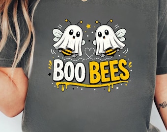 May include: A charcoal grey t-shirt featuring a graphic of two bees dressed as ghosts. The text "BOO BEES" is printed in large white and yellow letters below the bees, with a yellow dripping honey effect underneath the text and small stars.