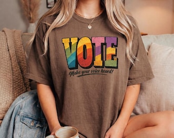 May include: A brown short-sleeve t-shirt featuring the word VOTE in large, colourful, rainbow-striped block letters with a distressed texture. Below the word VOTE, the text Make your voice heard! is printed in a black, cursive-style font.