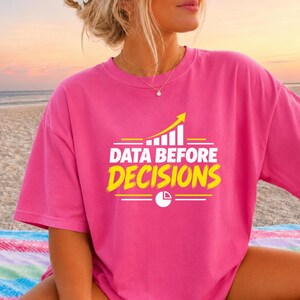 May include: A bright pink short-sleeved t-shirt featuring a yellow and white graphic print. The design includes a rising bar chart icon, the text "DATA BEFORE DECISIONS" in bold white and yellow lettering, and a small pie chart icon below.