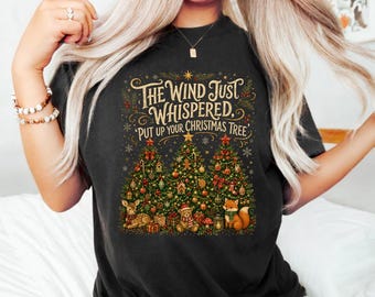 May include: A black t-shirt featuring a graphic of three decorated Christmas trees, a deer, a teddy bear, and a fox. The text reads, "The Wind Just Whispered, Put Up Your Christmas Tree," surrounded by gold stars, snowflakes, and festive greenery.