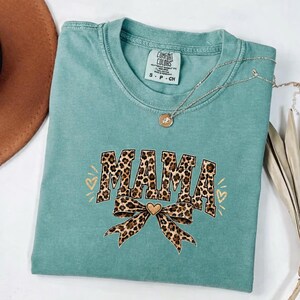 May include: A sage green short-sleeved t-shirt with a crew neckline. The front features the word MAMA in a brown and black leopard print pattern, accompanied by a matching leopard print bow graphic and small gold-coloured heart outlines.