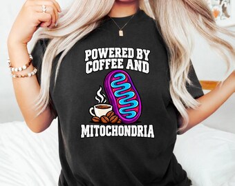 May include: A black t-shirt featuring a graphic print with the text "POWERED BY COFFEE AND MITOCHONDRIA". The design includes a white coffee cup with steam, a pile of brown coffee beans, and a purple and blue illustration of a mitochondrion.