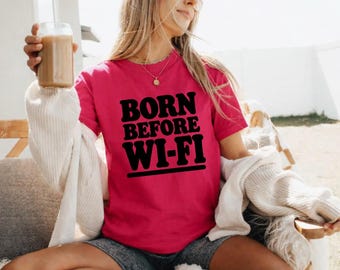 May include: A bright pink short-sleeved t-shirt featuring the black text BORN BEFORE WI-FI printed in a bold, retro-inspired font across the chest. The shirt is paired with grey shorts and a cream-coloured knit cardigan draped over the shoulders.