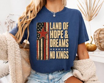 May include: A navy blue short-sleeve t-shirt featuring a distressed graphic of a vertical American flag with a red star and the text "LAND OF HOPE & DREAMS NO KINGS" in a bold, beige, block-style font with a row of small stars underneath.