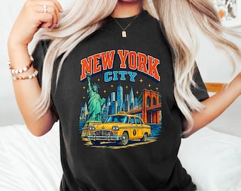 May include: A black t-shirt featuring a graphic print of the New York City skyline, the Statue of Liberty, the Brooklyn Bridge, and a yellow taxi cab. The text "NEW YORK CITY" is printed in large orange and blue letters above the illustration.