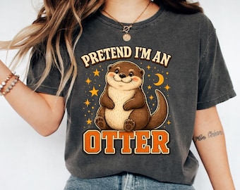 May include: A charcoal grey t-shirt featuring a graphic of a brown otter with pink cheeks, surrounded by yellow stars and a crescent moon. The text above reads "PRETEND I'M AN" and the text below reads "OTTER" in bold orange block letters.