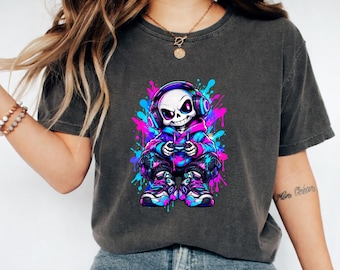 May include: A charcoal grey t-shirt featuring a graphic print of a skeleton wearing headphones and a hoodie while holding a game controller. The skeleton is surrounded by vibrant splashes of neon pink, blue, and purple paint, with matching trainers.