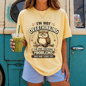 Könnte beinhalten: Ein hellgelbes Kurzarm-T-Shirt mit einer Grafik einer Eule auf einem Ast. Der Text lautet I'M NOT OVERTHINKING I'M PRE-SOLVING PROBLEMS THAT DON'T EXIST YET. Das T-Shirt wird mit blau-weiß gestreiften Shorts mit vertikalen Streifen kombiniert.