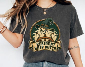 May include: A charcoal grey short-sleeve t-shirt featuring a graphic of a skeleton wearing a green witch hat and steering a ship's wheel. Below the illustration, a tan banner displays the text RESIDENT SHIP WITCH in bold black lettering.
