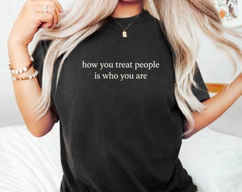 May include: A black short-sleeved t-shirt featuring the white text "how you treat people is who you are" printed in a simple font across the chest. The garment has a crew neckline and a standard fit, providing a clean and straightforward appearance.