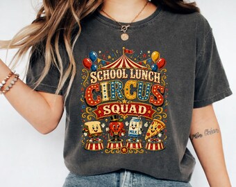 May include: A charcoal grey short-sleeve t-shirt featuring a colourful graphic print with the text "SCHOOL LUNCH CIRCUS SQUAD". The design includes a circus tent, balloons, and cartoon characters of a sandwich, an apple, a milk carton, and pizza.