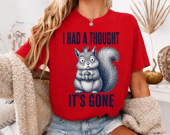 May include: A bright red short-sleeved t-shirt featuring a graphic of a grey squirrel holding an acorn. The text "I HAD A THOUGHT" is printed in dark blue above the squirrel, and "IT'S GONE" is printed in dark blue below the squirrel.